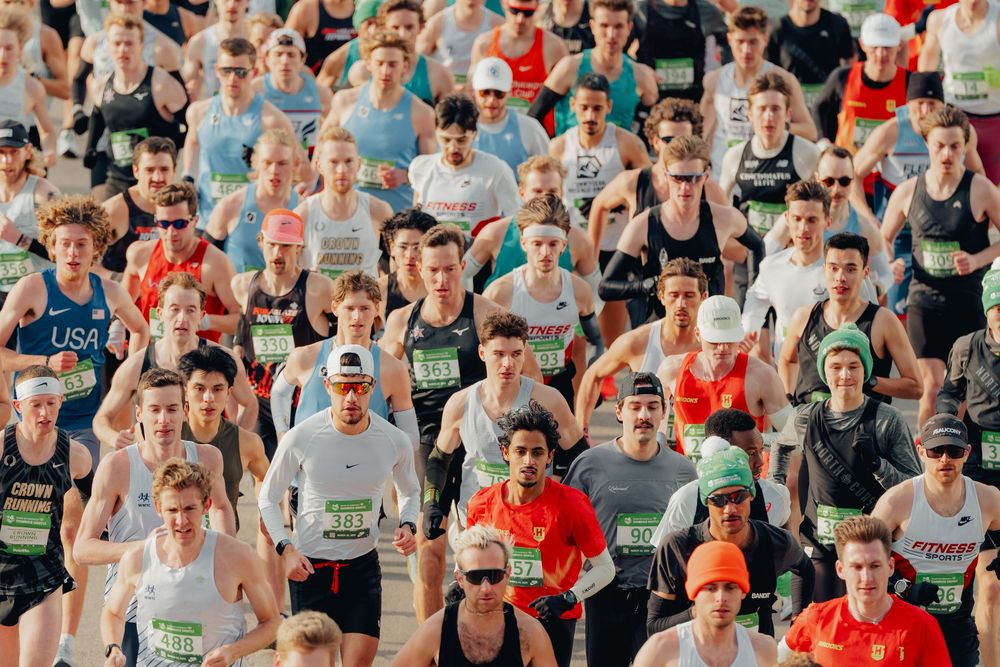 A large crowd of runners wearing race bibs compete in an outdoor marathon on a sunny day.