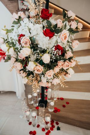 Elegant floral centerpiece with red and blush roses, white hydrangeas, and scattered rose petals on wooden table.