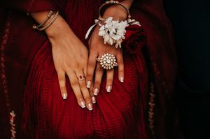 Close-up details of hands wearing a deep red dress with white floral accents and pearl jewelry.