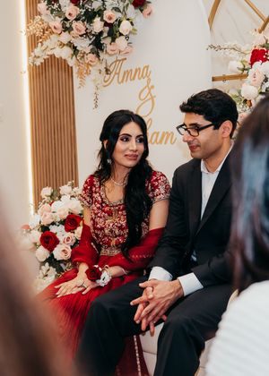 A couple in formal attire sits together on a decorated couch with red and pink floral arrangements in the background.