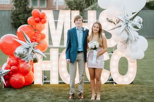 A couple stands in front of large HOCO letters decorated with red and white balloons and silver bows for homecoming.
