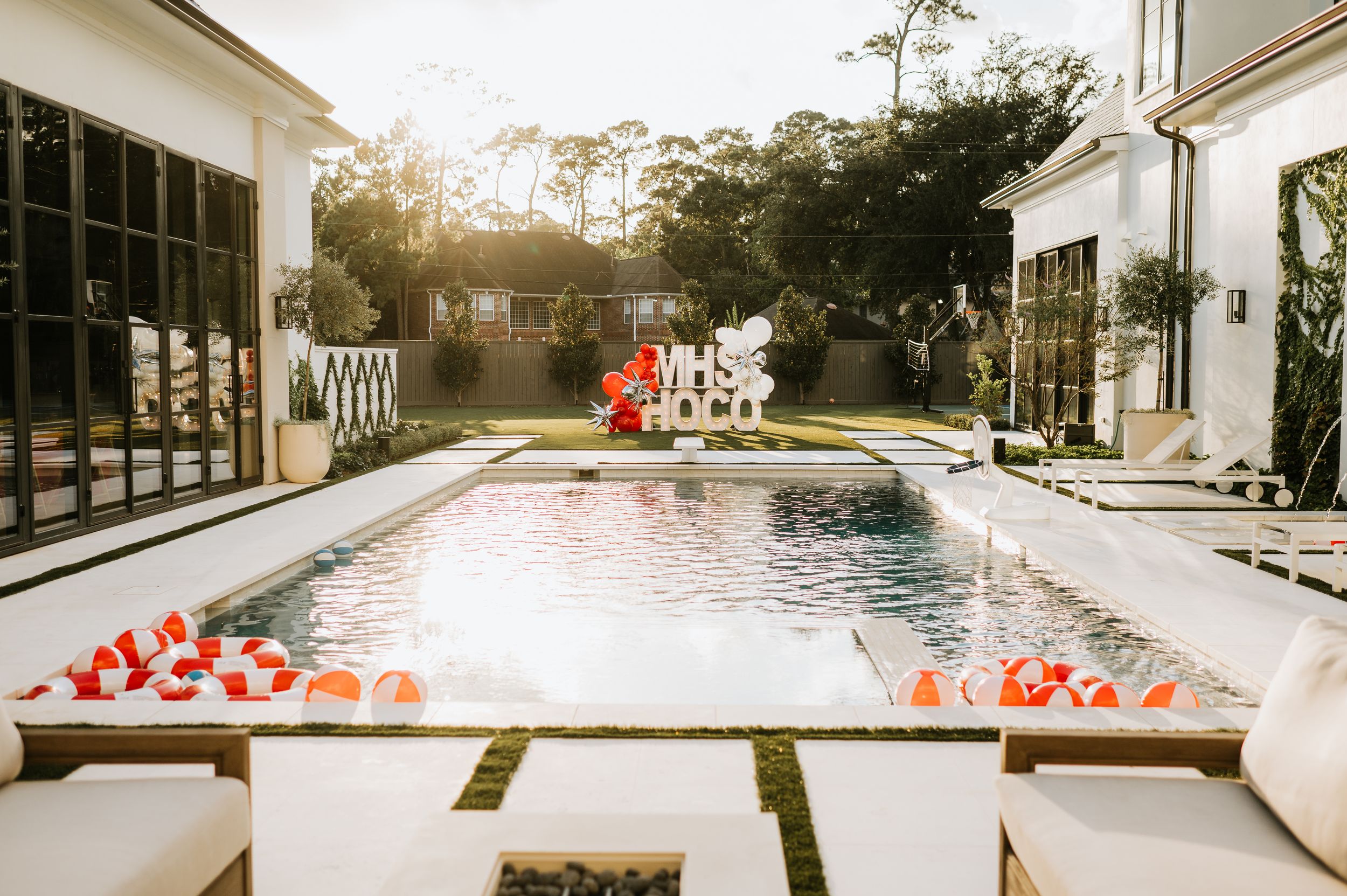 A modern backyard pool decorated with red and white heart-shaped balloons at sunset with palm trees in the background.