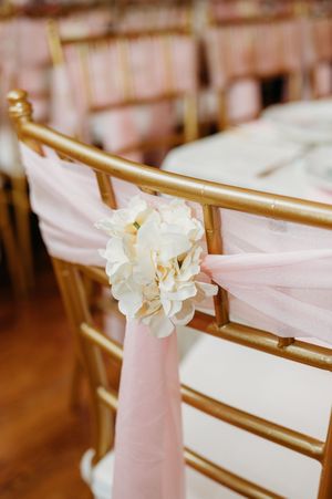Elegant wedding chair decorations featuring soft pink fabric sashes with white floral accents on gold chairs.
