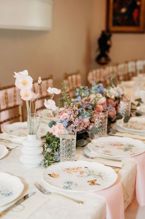 Elegant table settings with pink and blue floral centerpieces, crystal vases, and vintage china plates on white linens.
