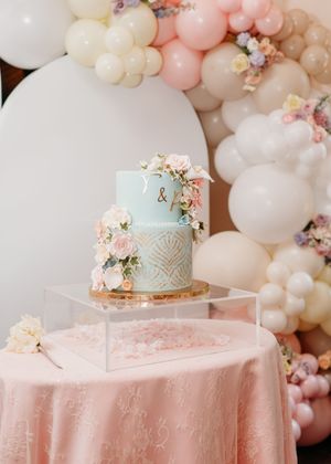 Elegant light blue and white wedding cake displayed on pink tablecloth with balloon arch decorations.