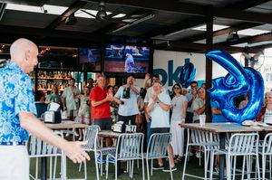 Patrons gather at an outdoor bar area with blue decor while watching sports on TV screens above.