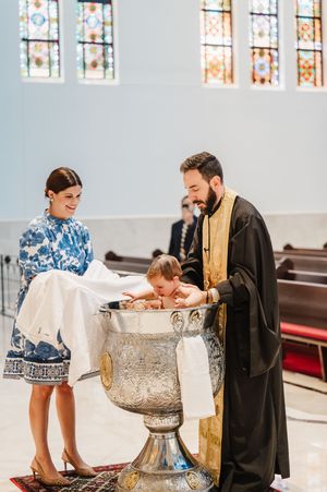 A religious baptism ceremony takes place at a church font with stained glass windows in the background.