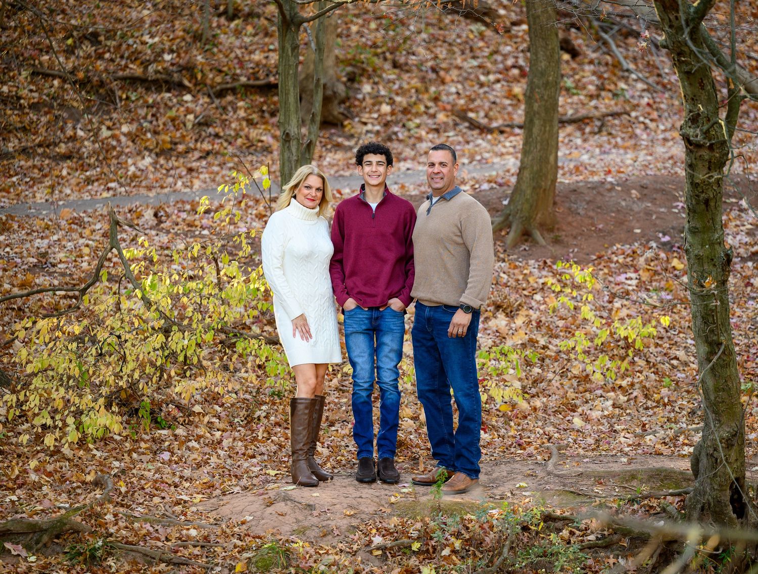 A family in coordinating blue and plaid outfits poses together on a wooded trail covered in fallen autumn leaves.