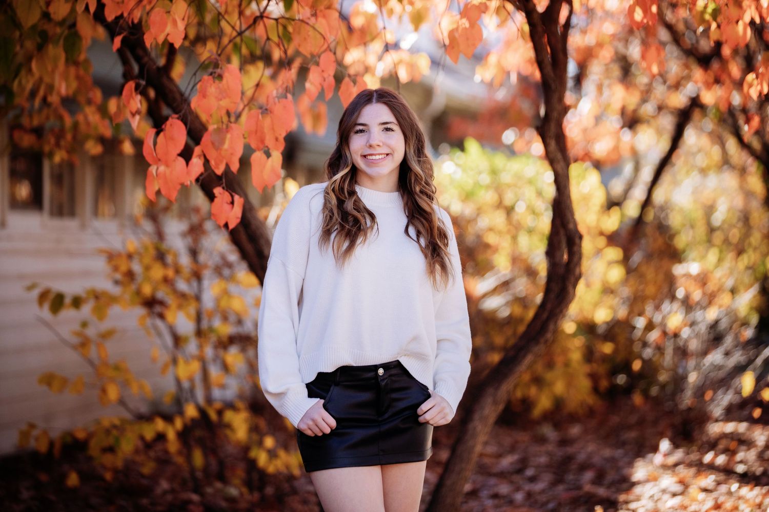 Fall senior portrait with white sweater against autumn foliage background.