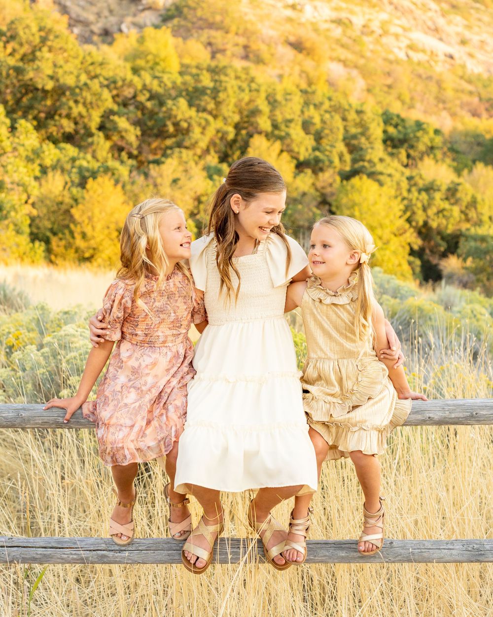 Three people in dresses sit on a wooden fence against a golden autumn hillside at hidden valley park, Utah.