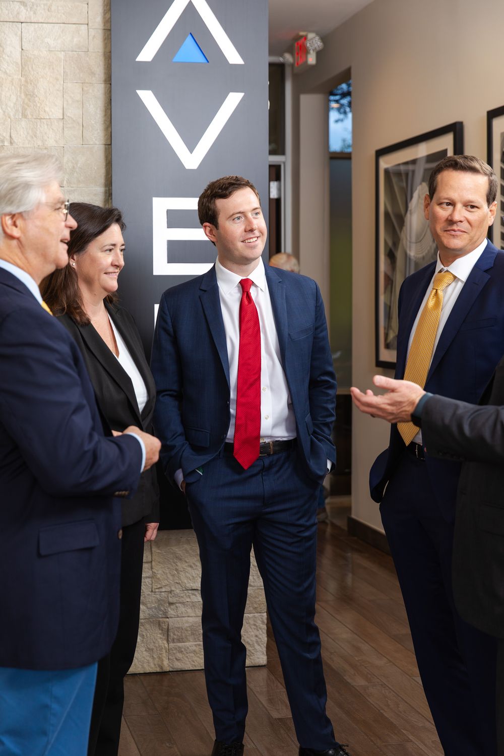 Four business professionals in suits conversing in a modern office lobby with a company logo on the wall.