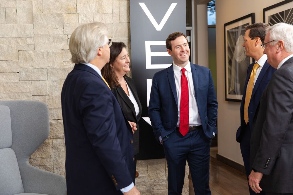 Business professionals in suits networking in a modern office lobby near a branded sign.