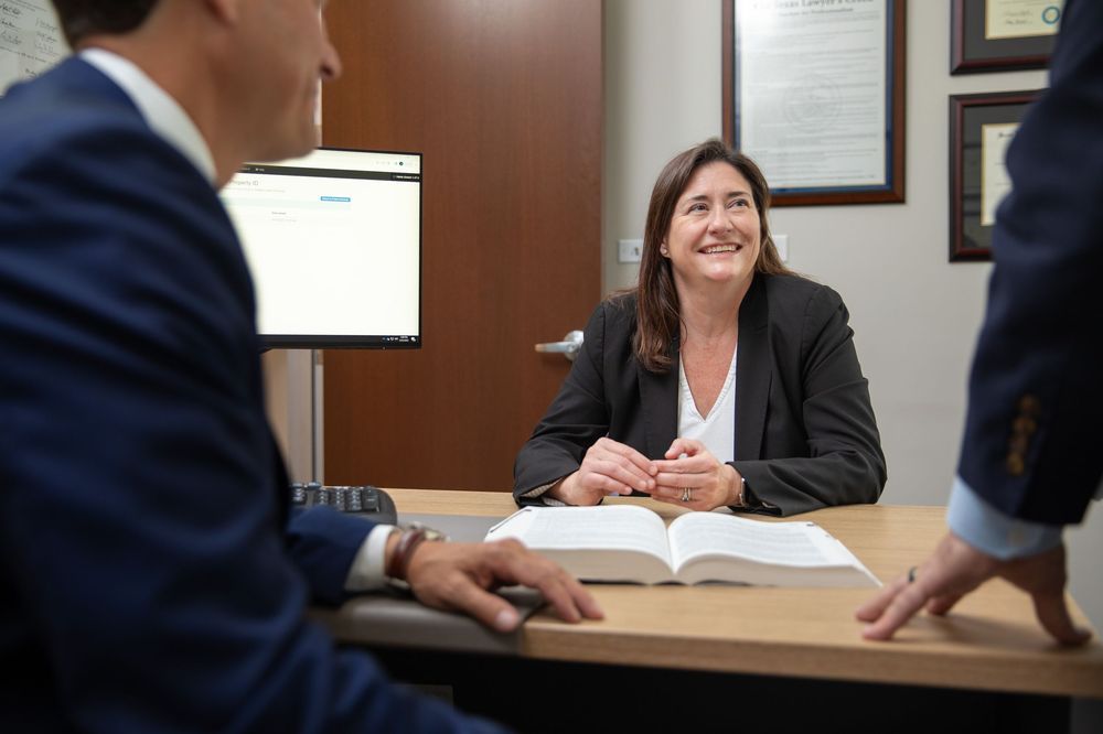 Professional woman in blazer smiling during business meeting at desk with open book and computer monitor.