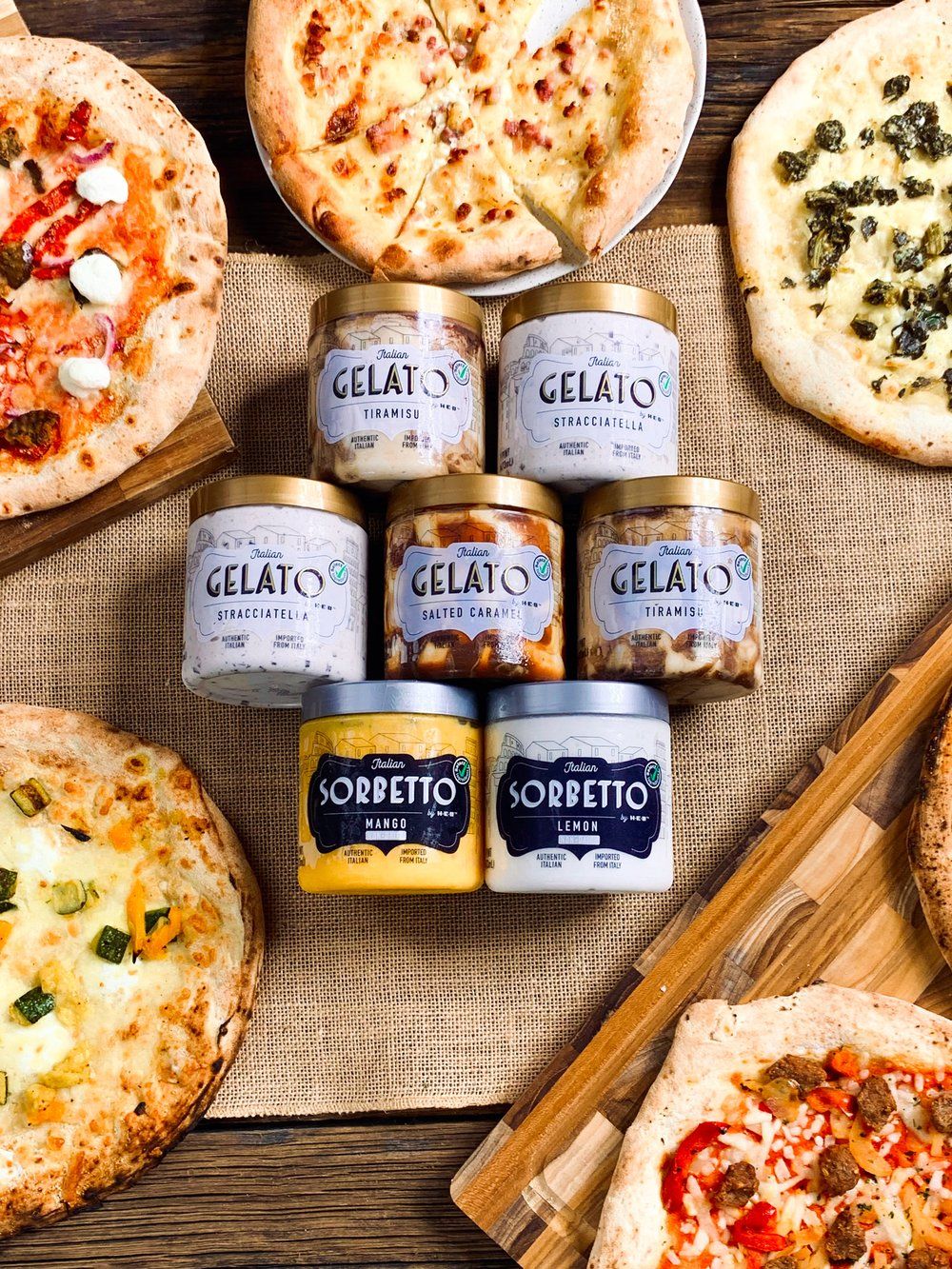 Flat lay of gourmet pizzas with Gelato branded condiment jars arranged on rustic surface.