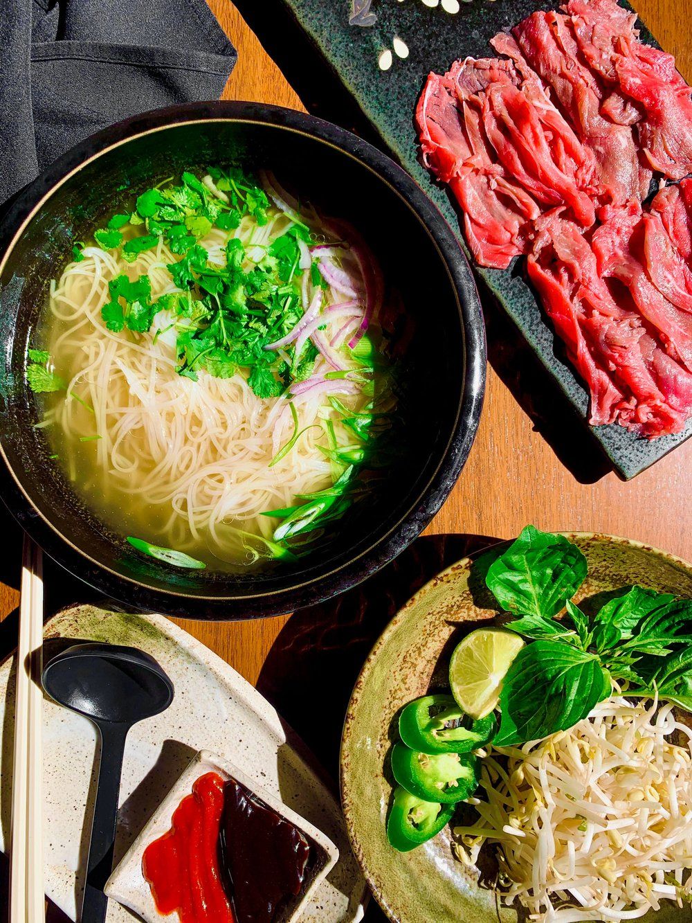 Fresh noodle soup preparation shown in stages with sliced meat, broth, and garnished with green herbs in black bowls.