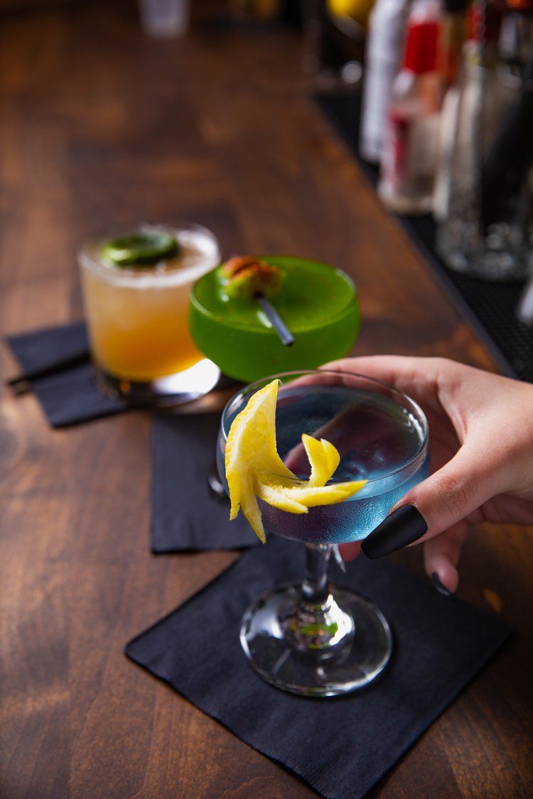 Colorful cocktails garnished with citrus twists served on dark coasters at a wooden bar.