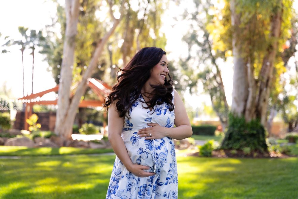 Smiling pregnant woman in floral dress cradling baby bump outdoors in lush green park setting.