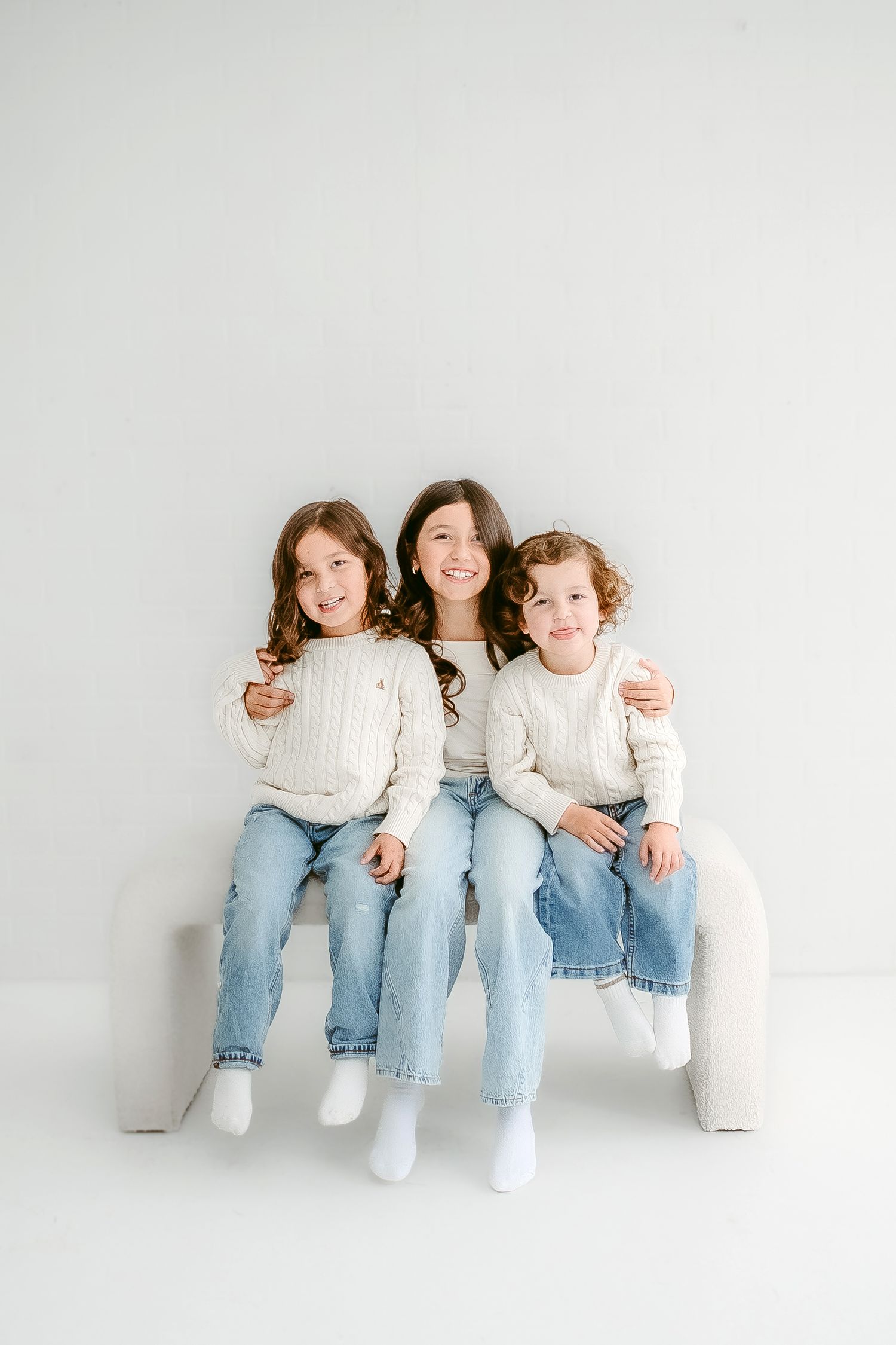 Three people sit together wearing matching white sweaters and blue jeans against a light gray background.