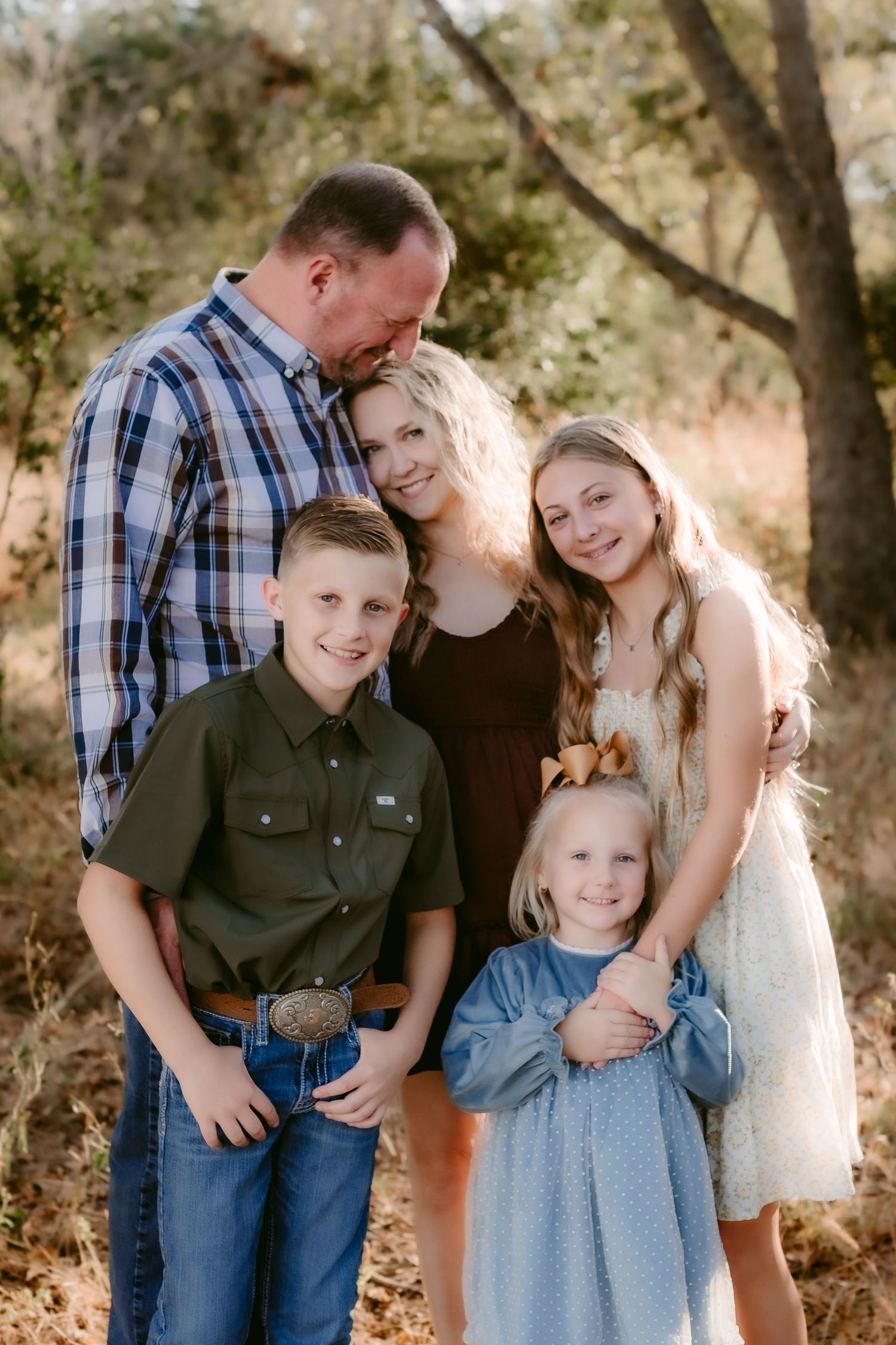 Family portrait session outdoors with natural lighting under trees showing candid loving moments in casual attire.