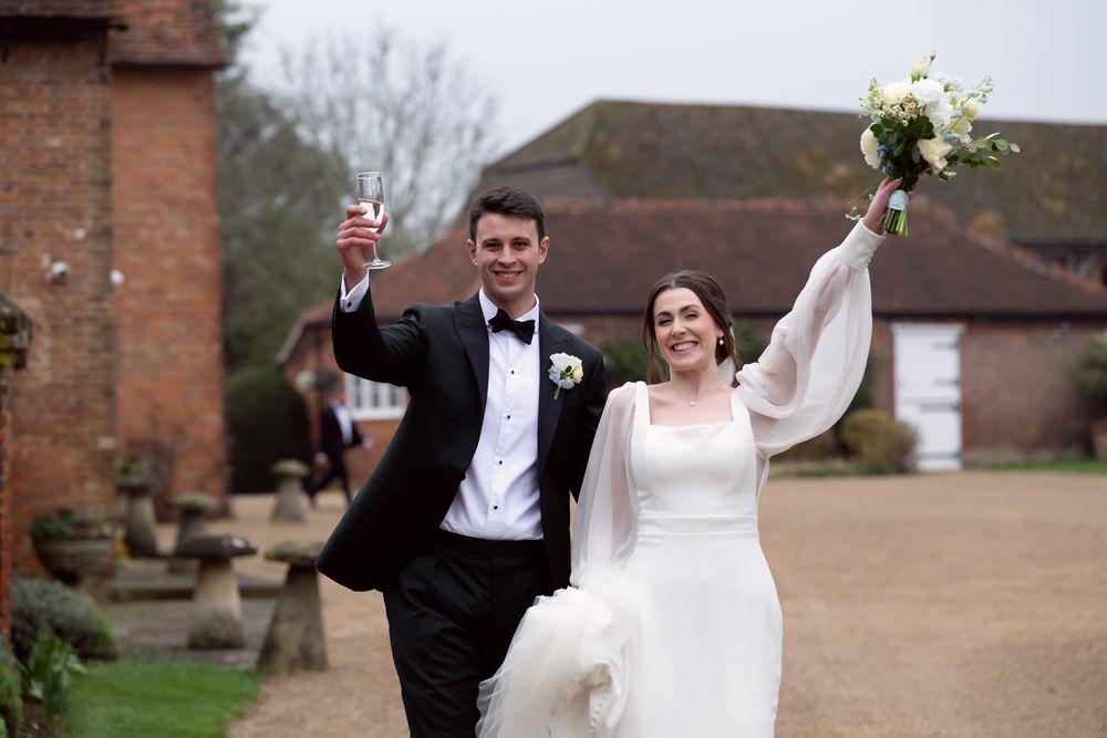 Joyful newlyweds celebrate outdoors, bride in white gown holding bouquet aloft, groom in tuxedo raising champagne glass.