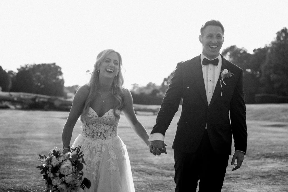 Black and white wedding photo of a couple walking hand in hand outdoors with the bride holding a floral bouquet.