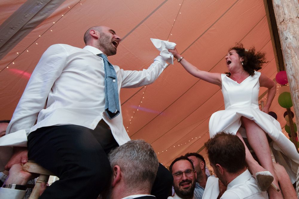 Bride and groom laughing joyfully while being lifted by guests during lively wedding reception celebration.