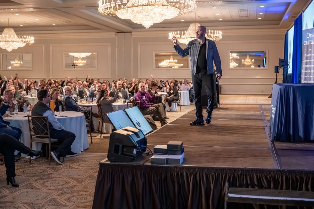 Speaker presenting on stage to a large audience in an elegant ballroom with chandeliers at a professional conference.