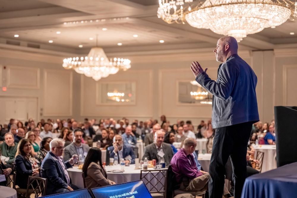 Speaker addresses large audience in elegant ballroom with crystal chandeliers at a professional conference event.