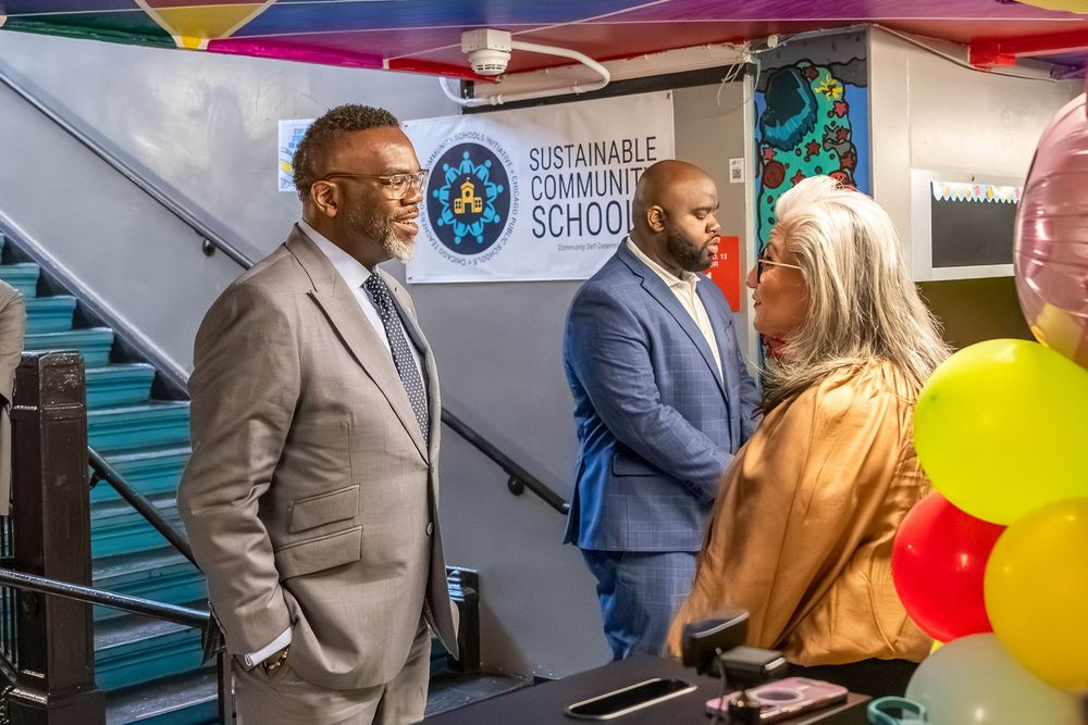 Three people conversing near a Sustainable Community School sign with colorful balloons at an indoor event.