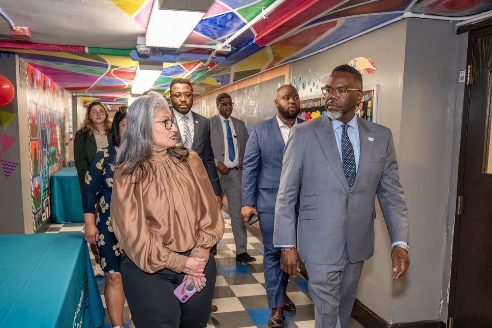 Officials in formal attire tour a vibrant, colorfully decorated community center hallway with rainbow streamers.