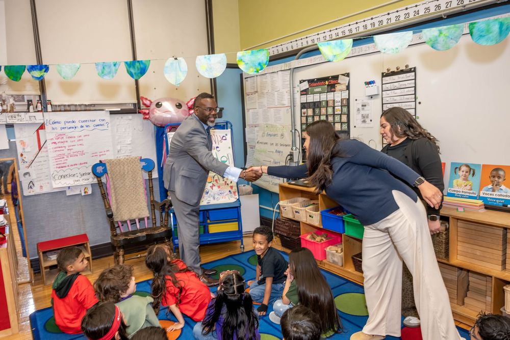 Adults engage with young students sitting on a colorful rug in a decorated elementary school classroom.