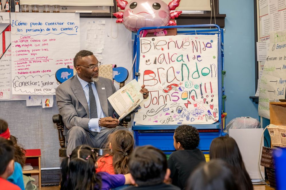 Man in suit reads a book to young students in a decorated classroom with a welcome sign.
