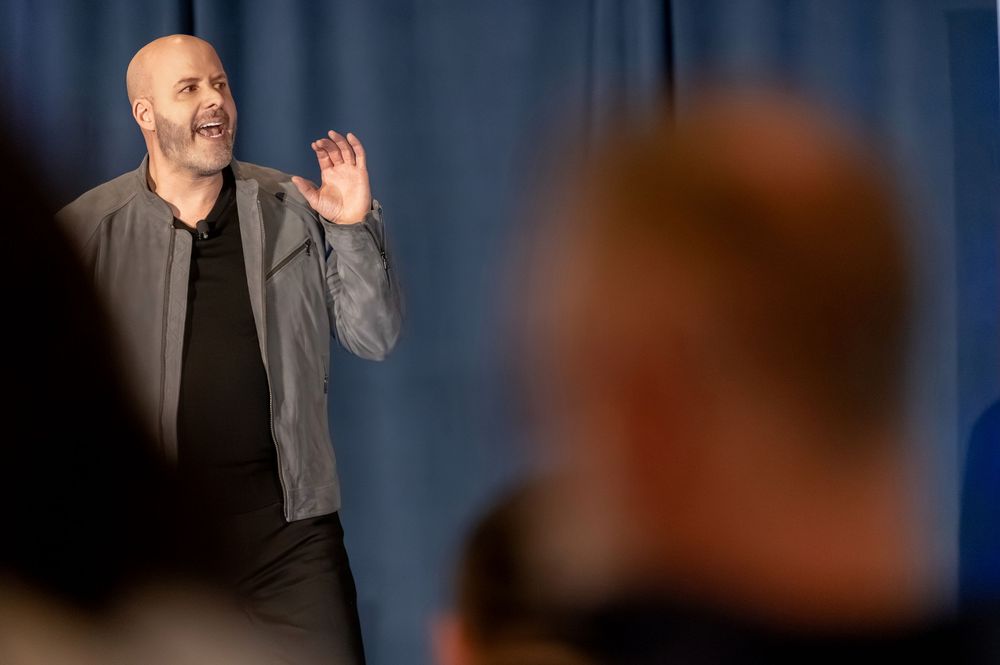 A bald man in a gray blazer gestures while speaking to an audience at a conference or seminar event.