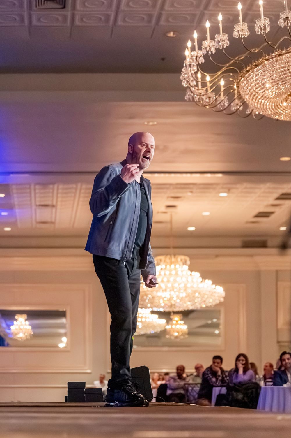 Bald man performing on stage in elegant ballroom with crystal chandeliers and seated audience in background.