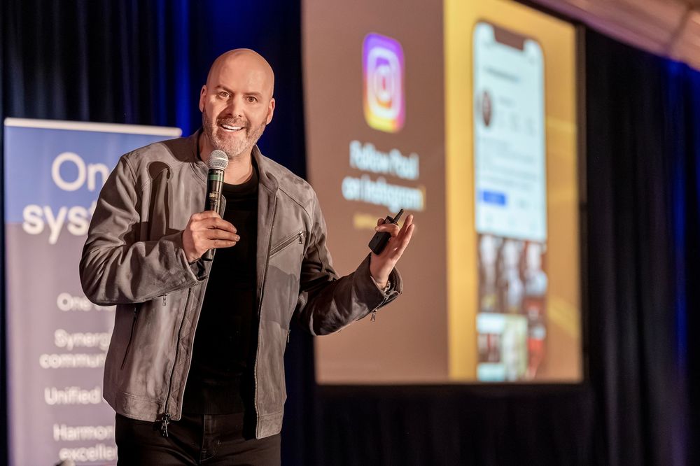 Bald man in gray jacket presents on stage holding microphone with Instagram screen displayed on projector behind him.