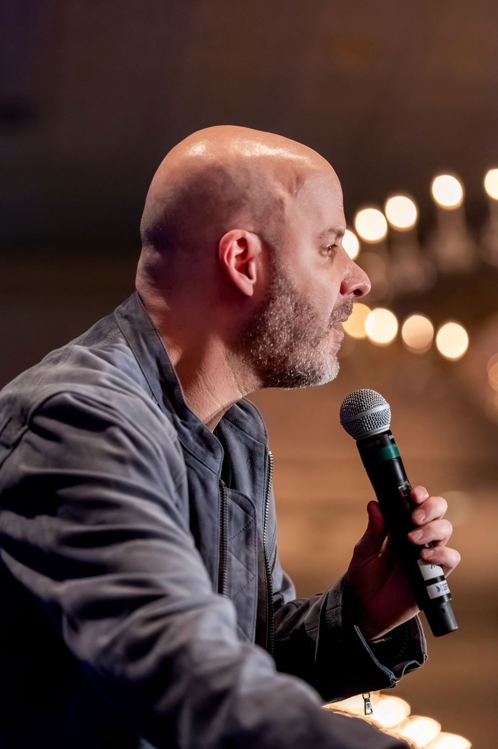 Bald man with beard speaking into a microphone on stage, with warm bokeh lights in the background.
