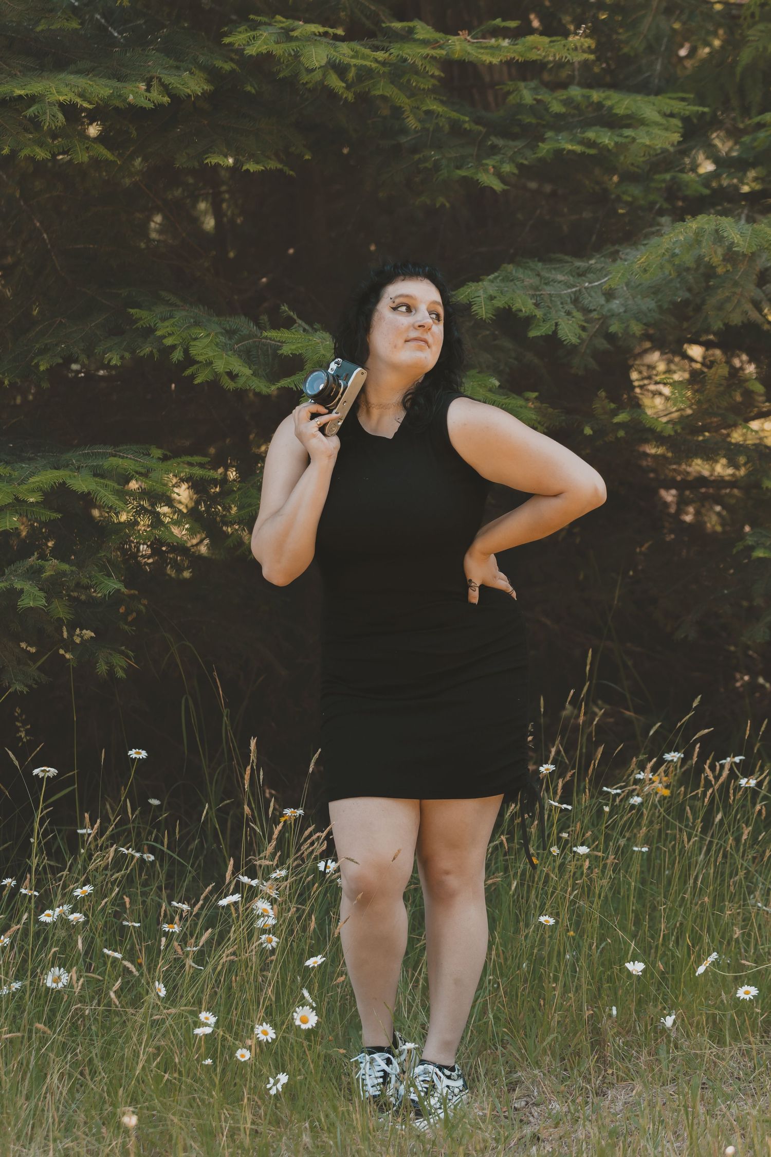 A woman standing in a field of daisys with a tree behind her. She is holding a camera. She is wearing checkered pants and a black shirt.