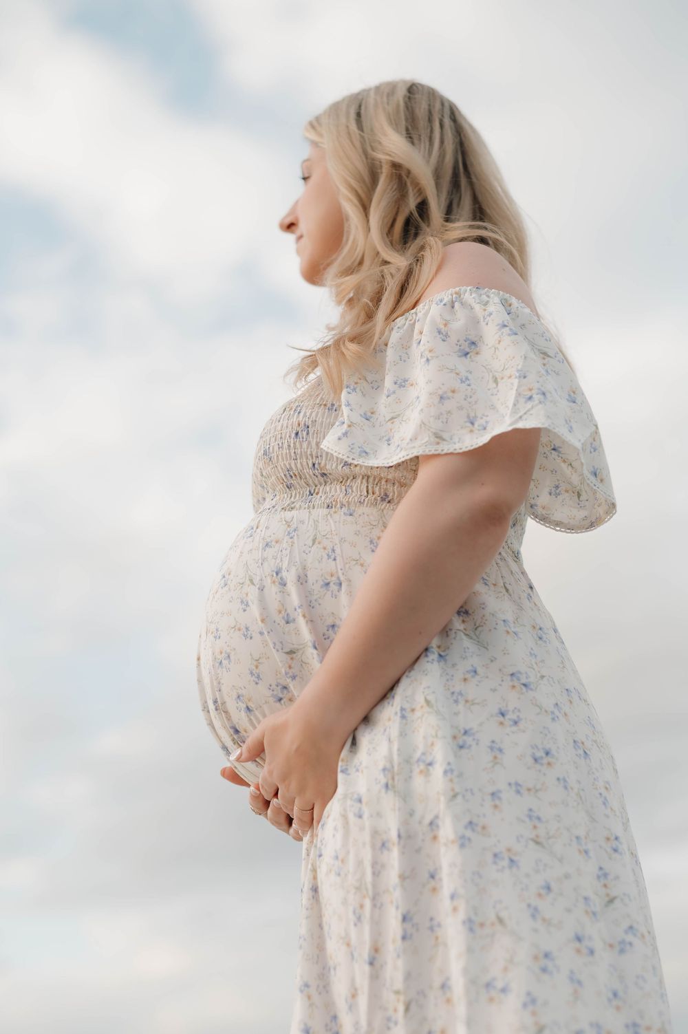 Pregnant woman in floral dress poses against cloudy sky showing baby bump profile.