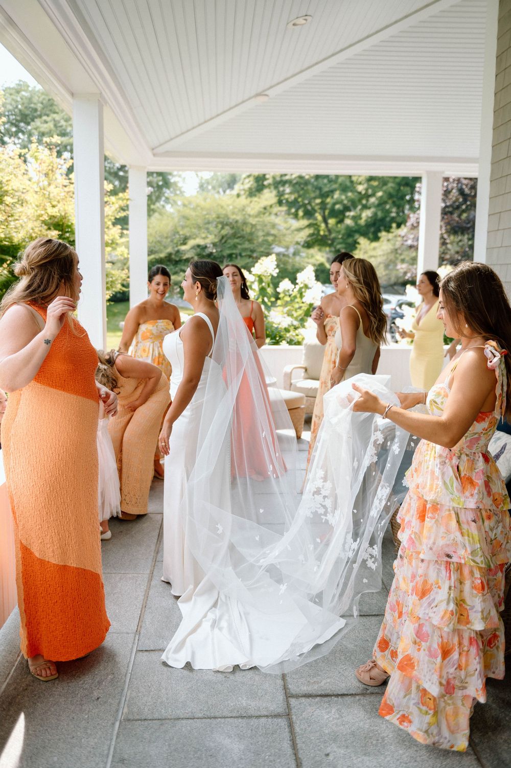 Wedding party in white and coral dresses gathered on covered porch for celebration photos.