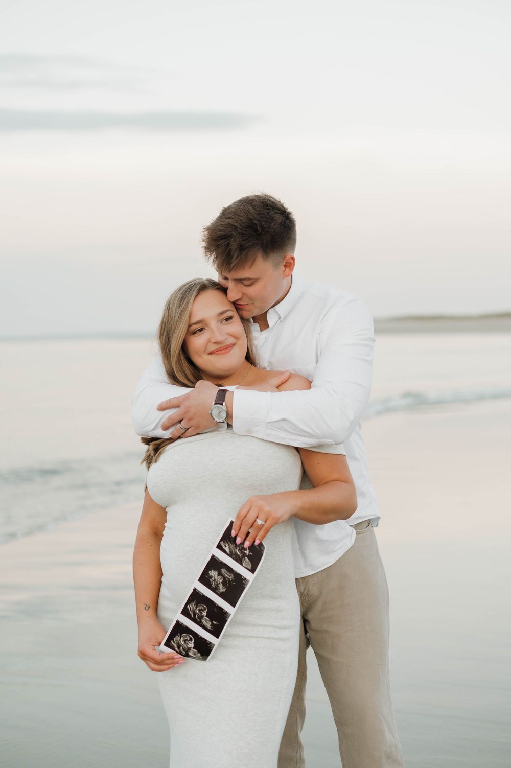 Couple embraces on beach holding ultrasound photos during sunset pregnancy announcement photoshoot.