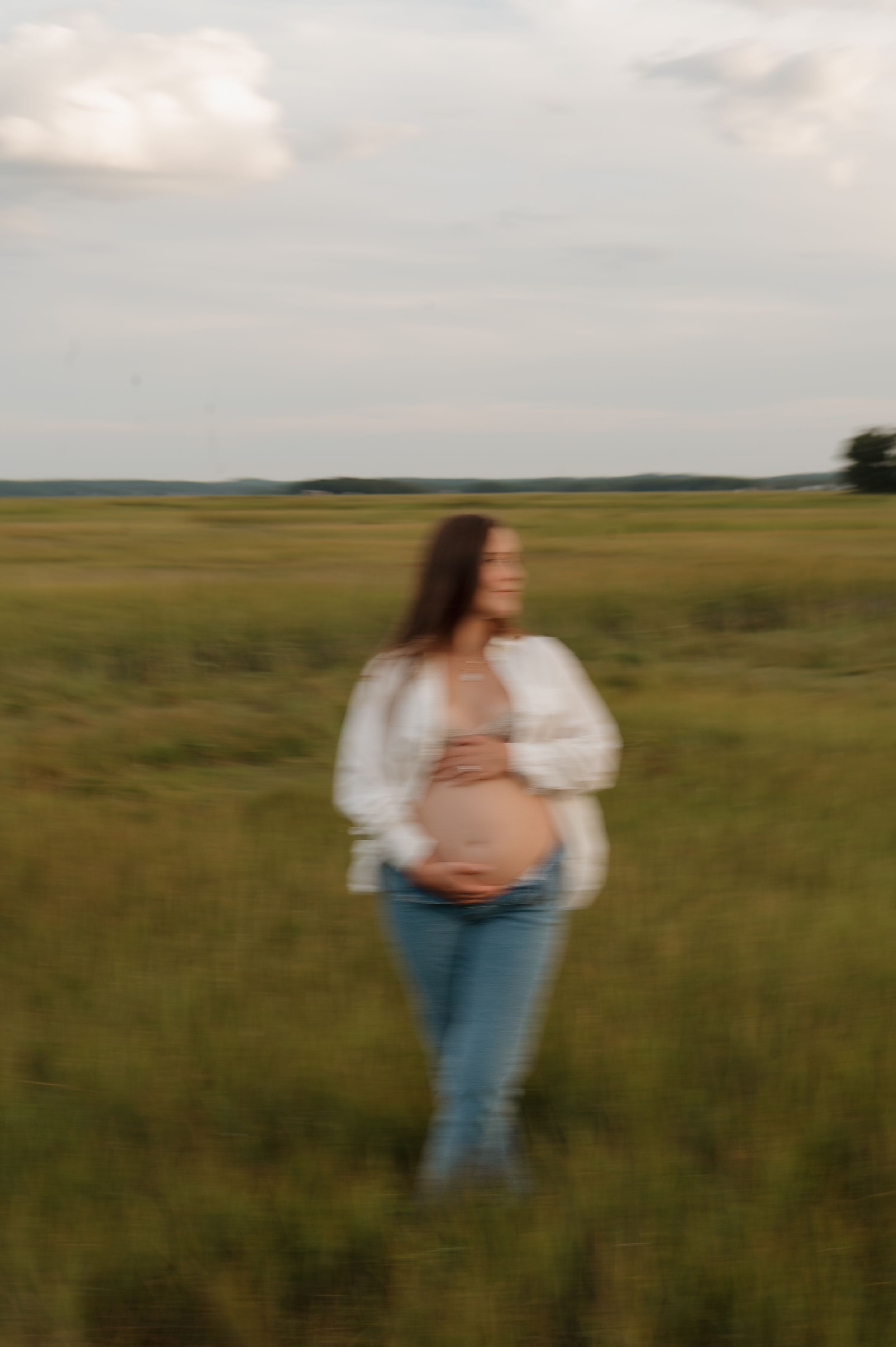 A figure stands in a serene prairie grassland wearing blue jeans and a white top against rolling hills and cloudy skies.