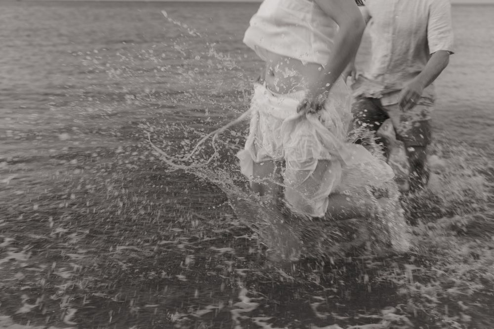 A couple wearing formal dress attire runs through ocean waves while celebrating and splashing in the water at sunset.