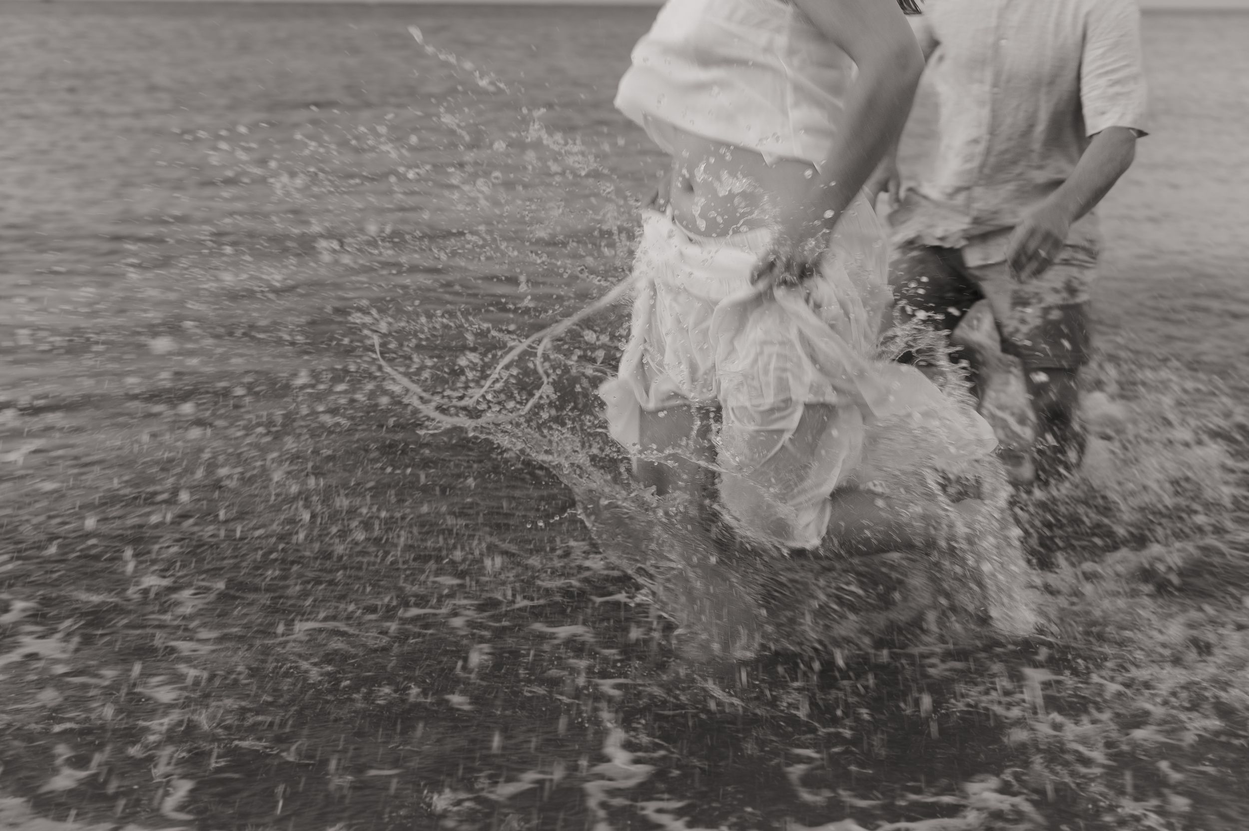 A couple wearing formal dress attire runs through ocean waves while celebrating and splashing in the water at sunset.