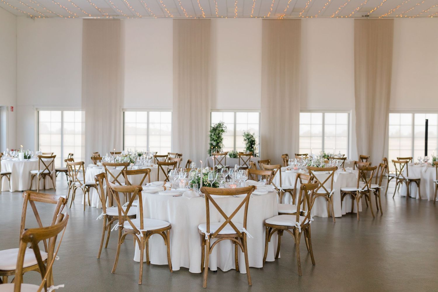 Elegant event space with white round tables, wooden cross-back chairs, and airy windows in a minimal modern setting.