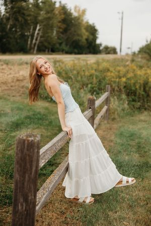 A person in a white summer dress twirls playfully in a sunny meadow next to a rustic wooden fence.