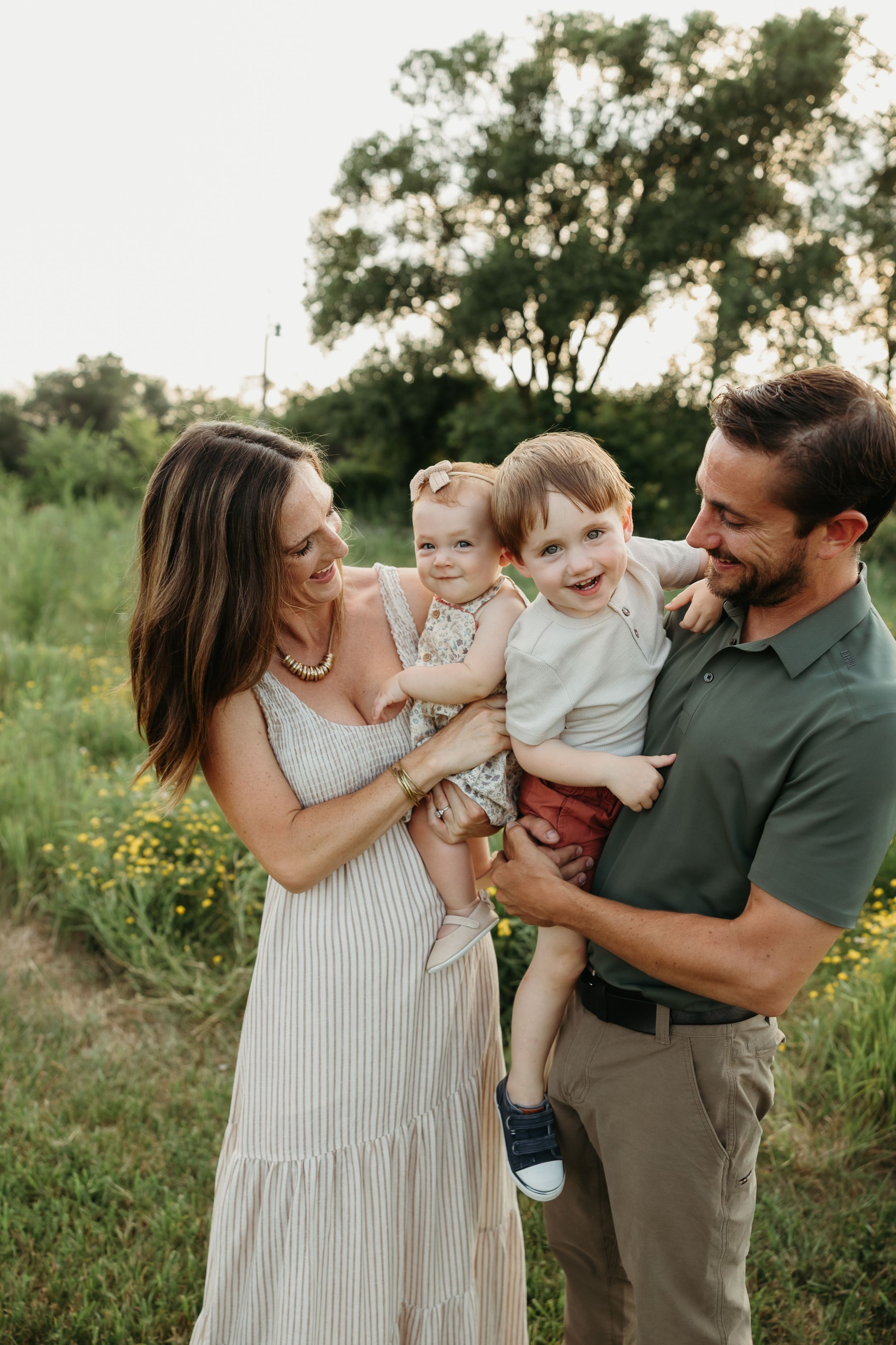 Family shares playful moments together outdoors in a natural setting with tall grass and wildflowers at sunset.