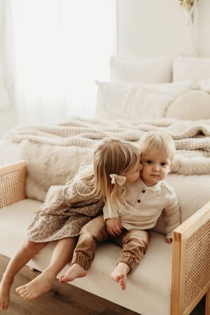 Two young children in neutral-toned clothing share a sweet moment together on a cane-sided daybed with cozy bedding.