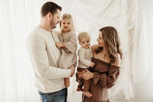 A family wearing neutral colors embraces while standing against a white curtain backdrop in a studio setting.