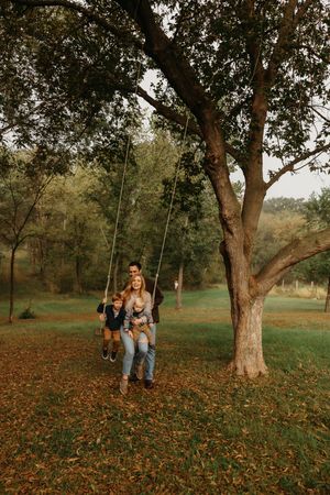 A couple sits together on a wooden tree swing in an autumn park surrounded by fallen leaves at sunset.