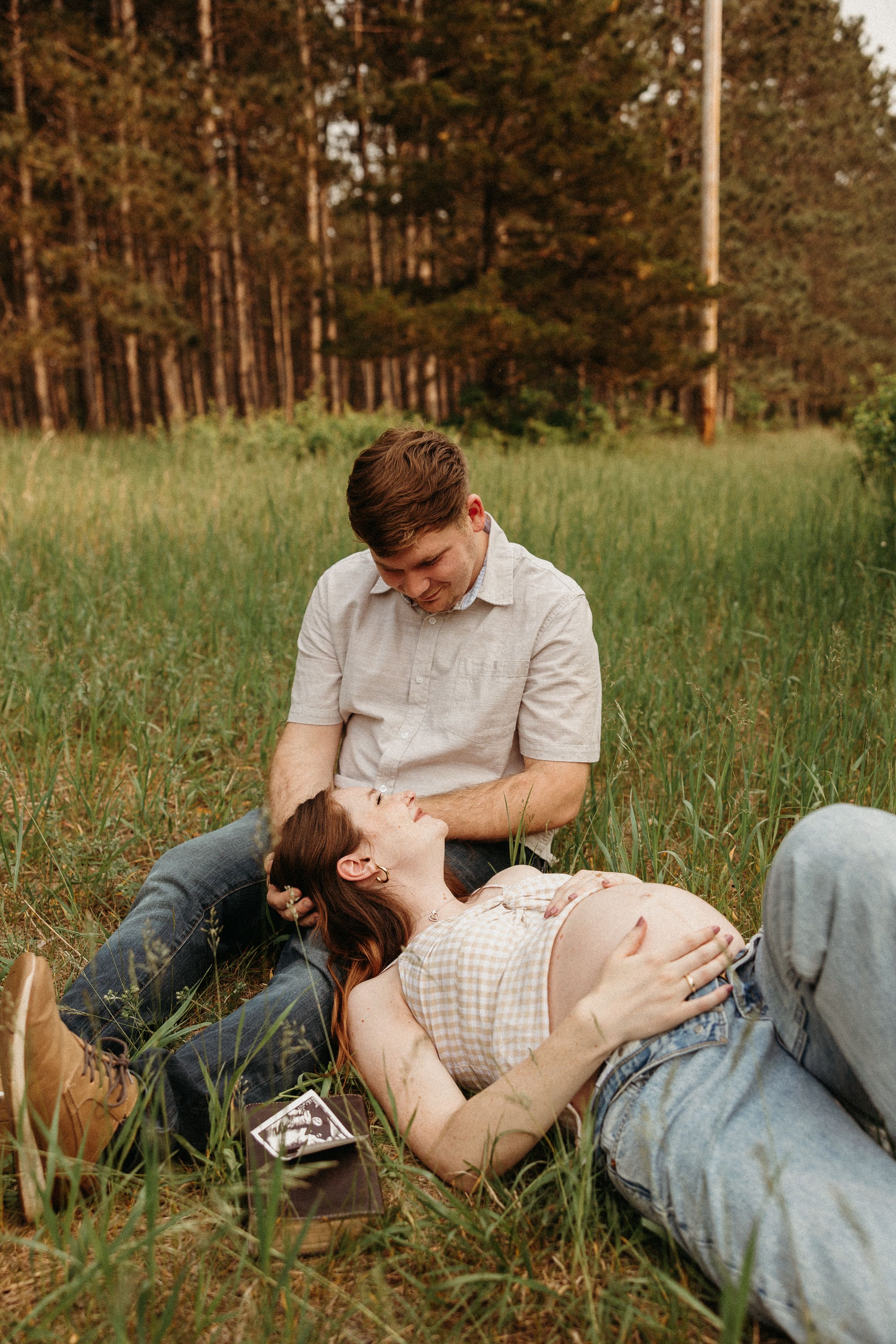 Couple lying together in tall grass field with pine trees in background during golden hour.